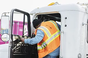 A man in an orange safety vest and black beanie climbing into the cab of a white semi-truck