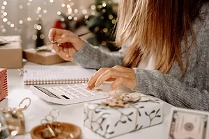 A woman in a grey sweater typing into a calculator next to a white notebook, and various wrapped gifts spread across the desk