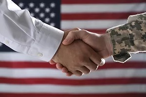 A handshake in front of an American flag between two men, one with a white sleeve and the other with military camo.