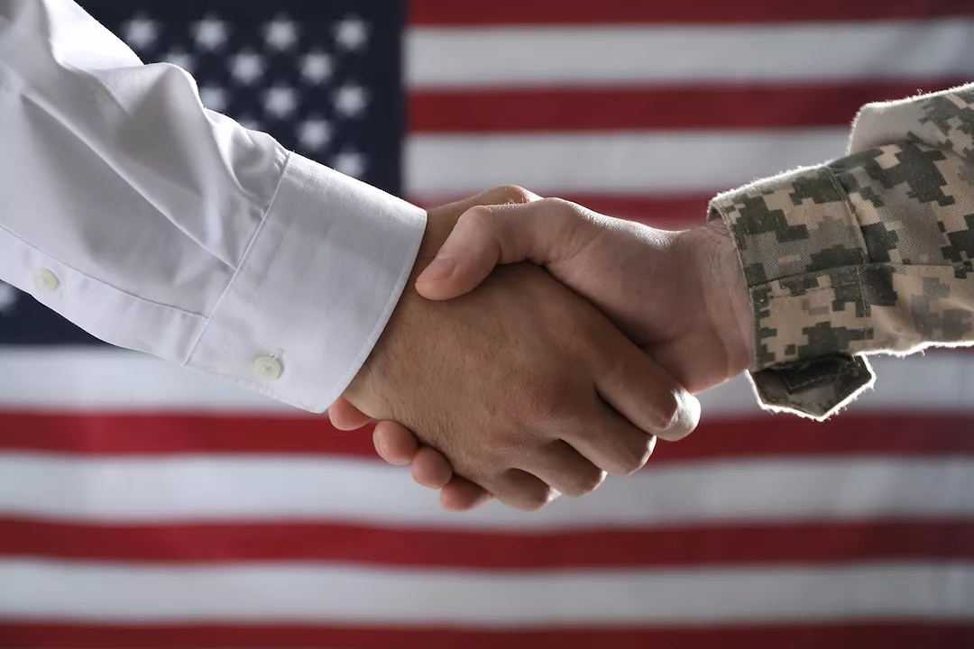 A handshake in front of an American flag between two men, one with a white sleeve and the other with military camo.