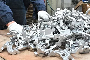 A pile of white, metallic gears and cogs, with a man in a blue jumpsuit and white gloves standing in front of the table.