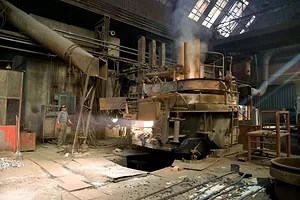 A WORKER IN BLUE OVERALLS AND A HARDHAT IS DWARFED BY A FURNACE IN A FACTORY.