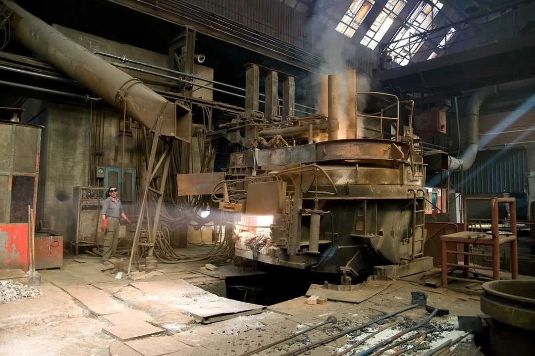A WORKER IN BLUE OVERALLS AND A HARDHAT IS DWARFED BY A FURNACE IN A FACTORY.