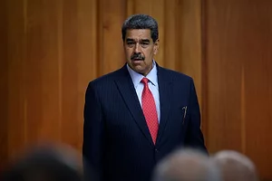 A MAN WITH GREYING HAIR AND MOUSTACHE, WEARING A BLUE SUIT WITH A RED TIE, ADDRESSES AN AUDIENCE.