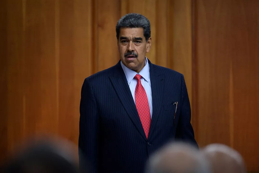 A MAN WITH GREYING HAIR AND MOUSTACHE, WEARING A BLUE SUIT WITH A RED TIE, ADDRESSES AN AUDIENCE.
