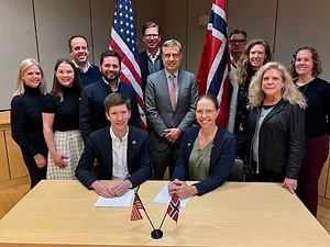 A GROUP OF PEOPLE CLUSTER AROUND A SIGNED DOCUMENT, FRAMED BY US AND NORWEGIAN FLAGS