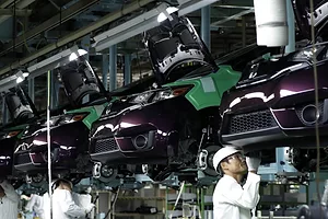 A line of cars hanging from hooks in a factory, with two men in white labcoats standing beneath the cars.