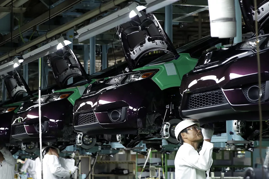 A line of cars hanging from hooks in a factory, with two men in white labcoats standing beneath the cars.