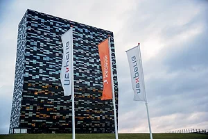 A tall glass office building with reflective blue windows, with three orange and white flags out front