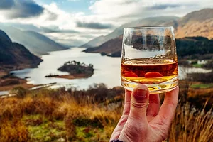A HAND HOLDS UP A GLASS OF GOLDEN LIQUID IN FRONT OF A DRAMATIC LAKE VALLEY WITH MOUNTAINS
