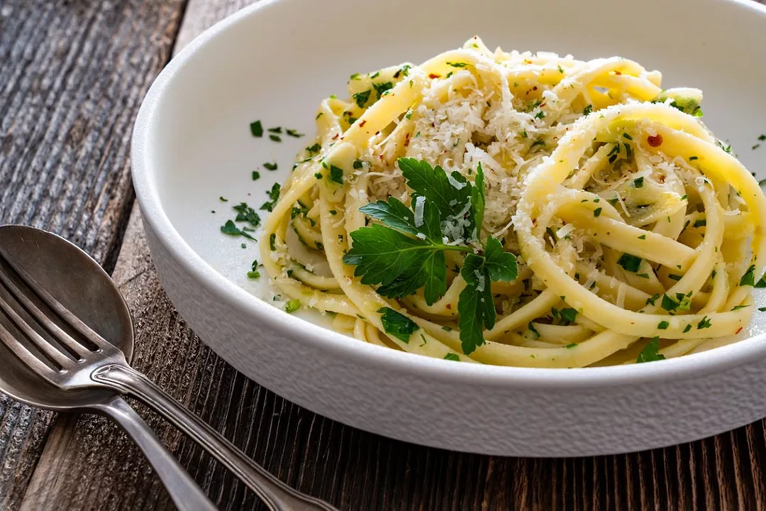 A bowl containing pasta with olive oil, parmesan and garlic sits on wooden table