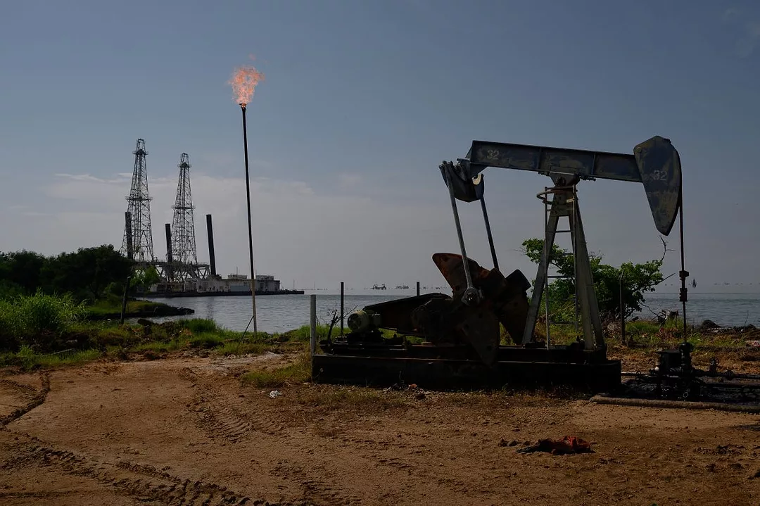 AN OIL JACK PUMPS IN THE FOREGROUND, WITH A REFINERY IN THE BACKGROUND
