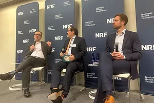 THREE MEN IN DARK SUITS AND WHITE SHIRTS SIT IN A ROW IN FRONT OF NRF BANNERS, ALL HOLDING MICROPHONES