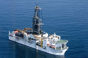 A white ship at sea with a tall scaffolded tower at its center, and a green helipad at the front.