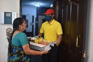 A MASKED DELIVERY DRIVER HANDS A BOX OF GROCERIES TO A WOMAN IN A SARI IN THE DOORWAY OF A HOME