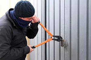 A MAN IN BEANIE AND FACE COVERING ATTACKS A CONTAINER SEAL WITH A PAIR OF BOLT CUTTERS