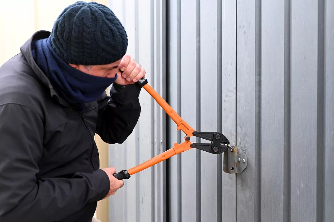 A MAN IN BEANIE AND FACE COVERING ATTACKS A CONTAINER SEAL WITH A PAIR OF BOLT CUTTERS