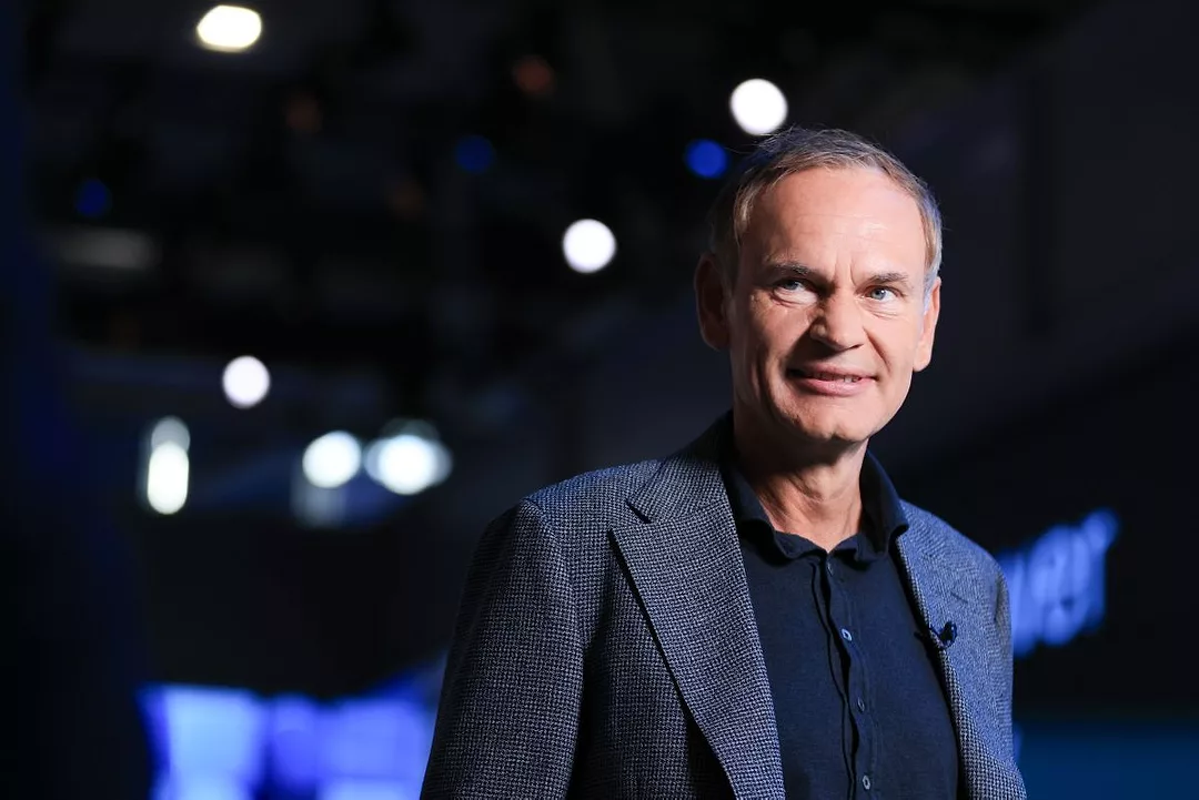 A MAN IN A JACKET AND SHIRT STANDS IN FRONT OF BLURRY LIGHTS