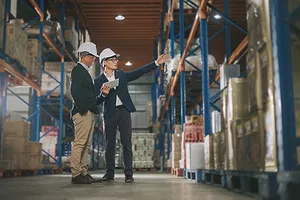 Two men in navy blue suits and white hard hats in a warehouse, with one holding a tablet and gesturing toward a large metallic blue shelf stacked with cardboard boxes
