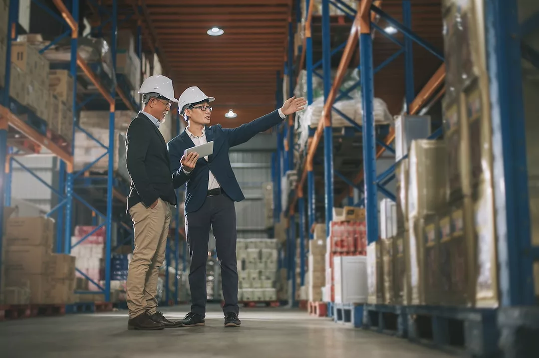 Two men in navy blue suits and white hard hats in a warehouse, with one holding a tablet and gesturing toward a large metallic blue shelf stacked with cardboard boxes