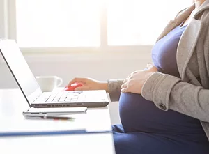 A PREGNANT WOMAN SITS TYPING ON A LAPTOP