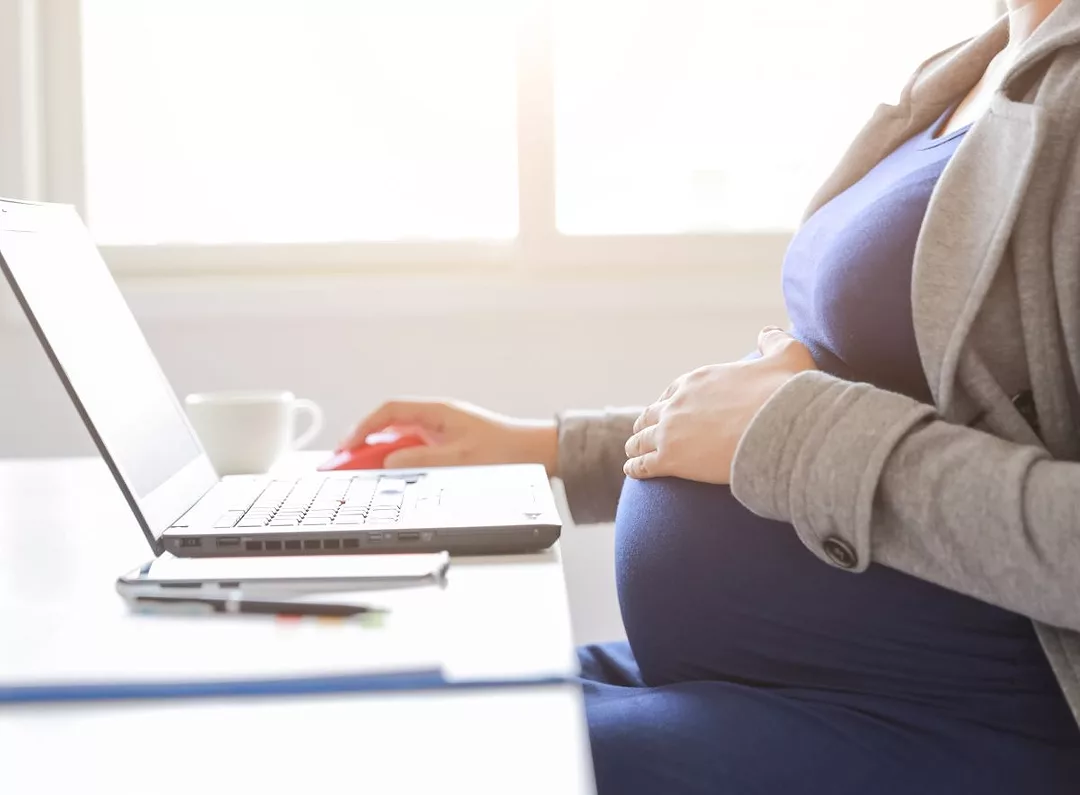 A PREGNANT WOMAN SITS TYPING ON A LAPTOP
