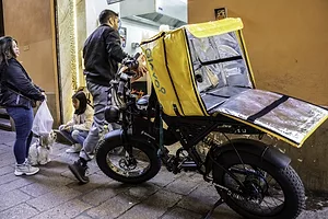 A man in a black leather jacket and jeans delivering food in front of a black motorcycle with a large yellow cooler case that reads "Glovo" on the side in blue lettering