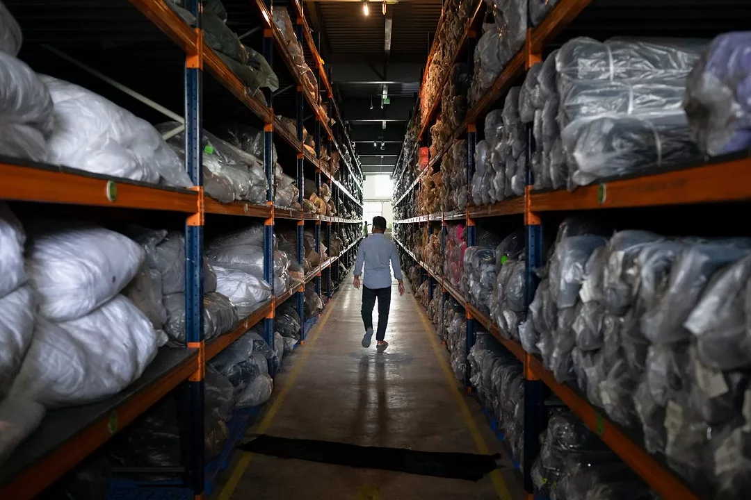 A MAN WALKS DOWN A LONG ROW OF SHELVES HOLDING BALES OF FABRIC