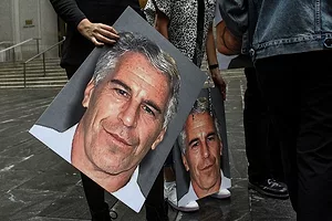 TWO PEOPLE SEEN FROM THE WAIST DOWN HOLD GIANT PICTURES OF A MAN