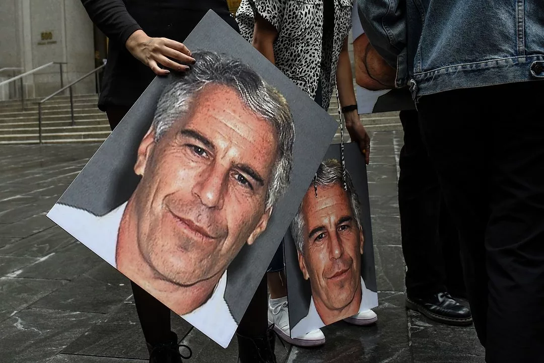 TWO PEOPLE SEEN FROM THE WAIST DOWN HOLD GIANT PICTURES OF A MAN