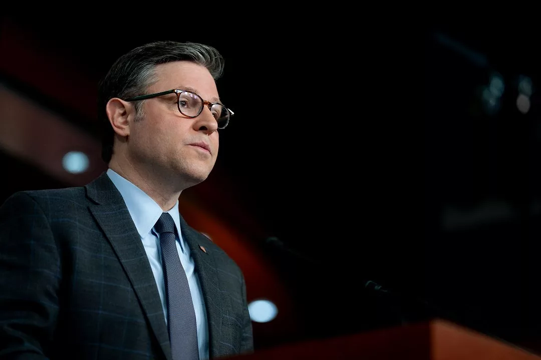 A MAN IN A BLUE SUIT AND SPECTACLES APPEARS TO PAUSE FOR THOUGHT DURING PUBLIC SPEAKING