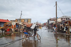 A man rides a red bicycle across a flooded street with damaged homes lining either side of the roadway