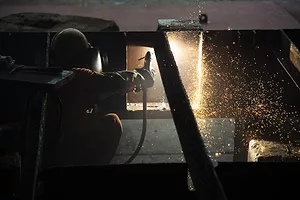 A person in a welding mask operating a blowtorch in front of a large steel panel