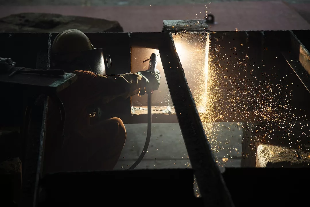 A person in a welding mask operating a blowtorch in front of a large steel panel