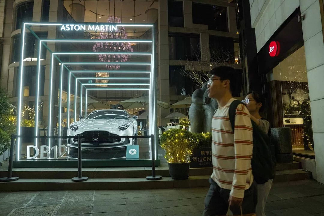 A YOUNG PERSON WEARING A BACKPACK WALKS PAST A FANCY LIT WINDOW DISPLAY OF A LUXURY CAR