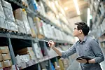Young Asian man worker doing stocktaking of product in cardboard box on shelves in warehouse by using digital tablet and pen.