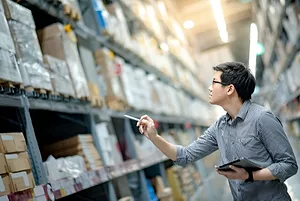 Young Asian man worker doing stocktaking of product in cardboard box on shelves in warehouse by using digital tablet and pen.