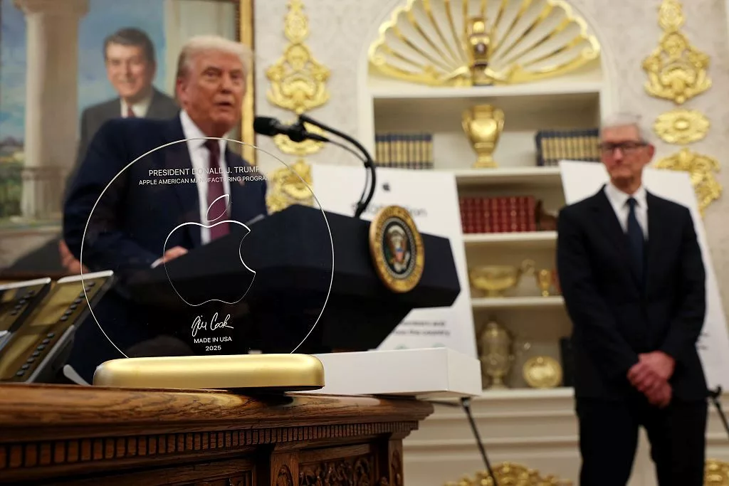 A MAN IN A SUIT SPEAKS AT A PODIUM IN A GILDED ROOM, WITH ANOTHER MAN IN A SUIT STANDING NEARBY
