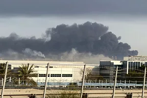 A HUGE PLUME OF BLACK SMOKE RISES OVER BUILDINGS, A CHAIN LINK FENCE IN THE FOREGROUND