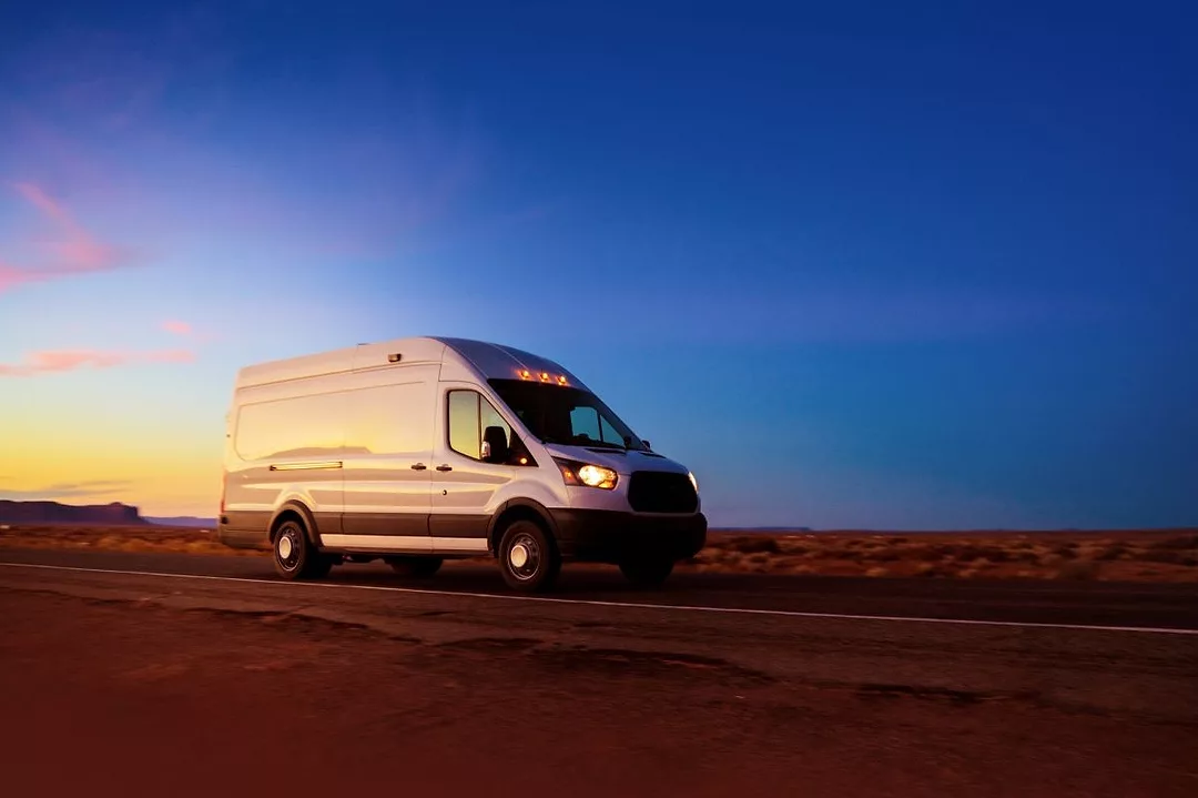 A WHITE DELIVERY VAN DRIVES DOWN A DESERT ROAD AT SUNSET