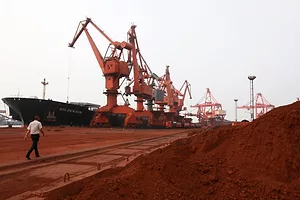 A MAN WALKS THROUGH A DOCK WITH A SHIP UNDER CRANES NEXT TO A PILE OF ORANGE RAW MATERIAL