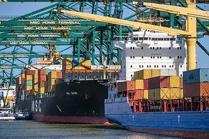 A black container ship docked underneath a row of blue shipping cranes