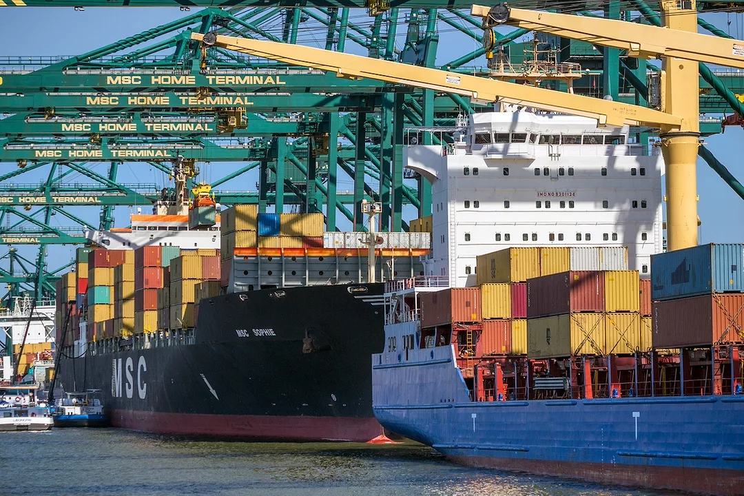 A black container ship docked underneath a row of blue shipping cranes