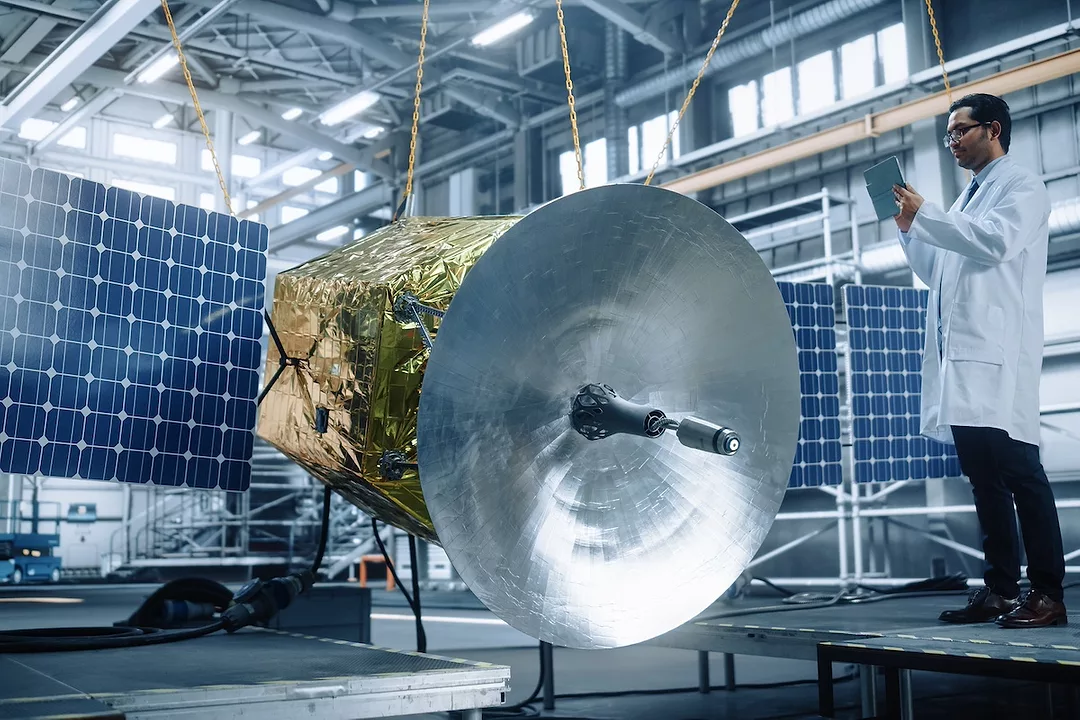 A man in a white labcoat stands next to a space satellite with a large white dish at its center, a gold foil body and a solar panel attached to either side