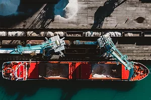An overhead view of a red container ship docked at a port, as a crane loads white powdered fertilizer onto the vessel