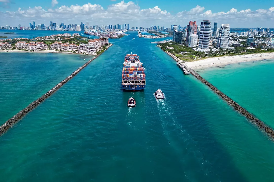 A container ship moving through a narrow waterway that feeds into a cityscape on a sunny day