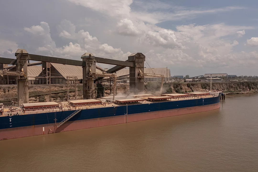 A long blue and red container ship being loaded with grain at a port on a cloudy day