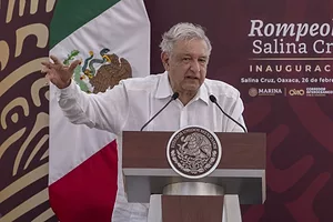 A GRAY-HAIRED MAN IN A WHITE SHIRT SPEAKS AT A PODIUM WITH FLAGS BEHIND HIM