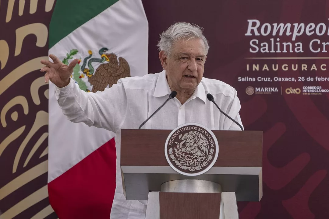 A GRAY-HAIRED MAN IN A WHITE SHIRT SPEAKS AT A PODIUM WITH FLAGS BEHIND HIM