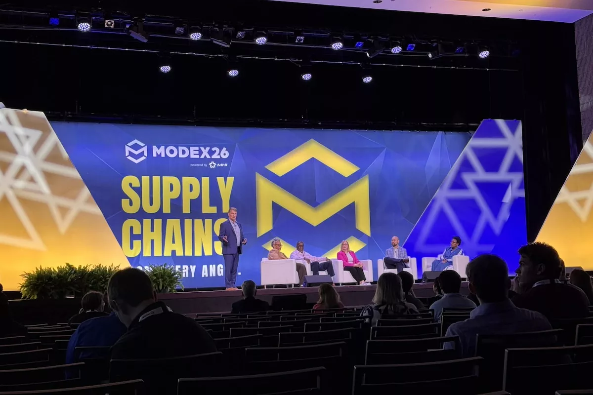 Takım elbiseli bir adam, arka planda "SUPPLY CHAINS" yazan büyük bir video ekranının önündeki sahnede dört dinleyiciye hitap ediyor